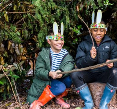 A photo of two young boys sitting on a log. They are smiling as they play with long twigs, and they are wearing rabbit ears attached to a paper crown.