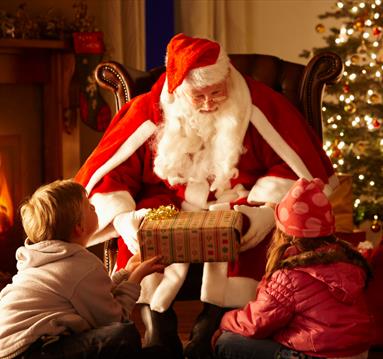 Two children being given a present by Santa in front of a Christmas tree and log fire