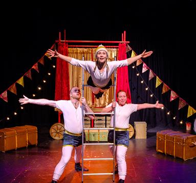 The image shows three people on stage, two men are holding a ladder from either side at the bottom of it. There is a woman with her waist on the ladde