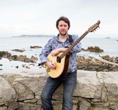 Irish singer and bouzouki player Daoirí Farrell sitting on a stone wall in front of a coastal scene