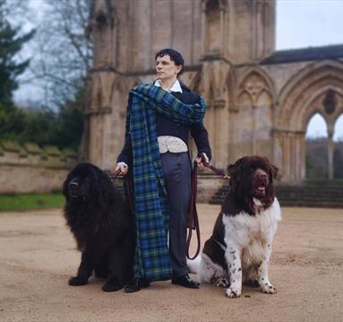 An actor dressed as Lord Byron poses in front of Newstead Abbey with two large Newfoundland dogs.