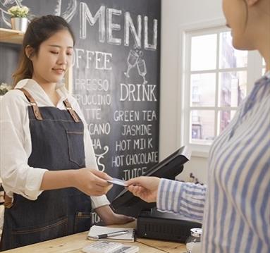 The image shows a lady behind a till taking something from someone's hand on the other side.
