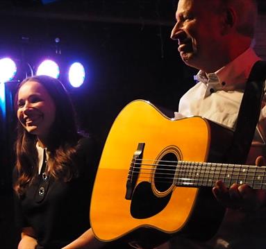 Image shows a man stood on stage playing a guitar and a woman stood next to him smiling.