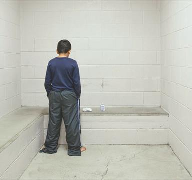 Photo from the exhibition showing a young person chained, facing a blank wall