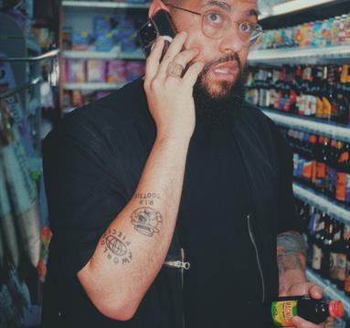 Jamali Maddix: Aston - Image shows a man on the phone stood in a supermarket wearing a black jumper and t shirt and holding two bottles of alcohol.