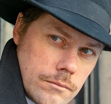 Man with fedora hat, pencil moustache and sinister appearance looks around a corner