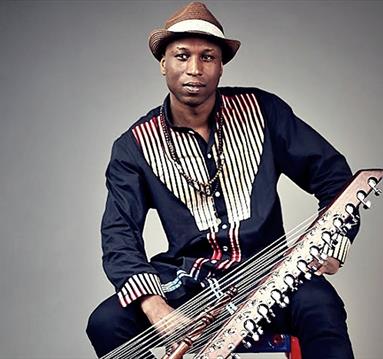 Image shows a man wearing a dark blue top with black and white stripes sat on a chair and holding an instrument.