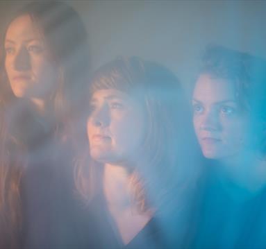 The image shows three women looking into the distance behind blue smoke.