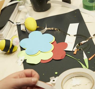 Image shows a craft table with coloured pieces of paper on it in the shape of flowers and a hand pulling some tape off a roll.