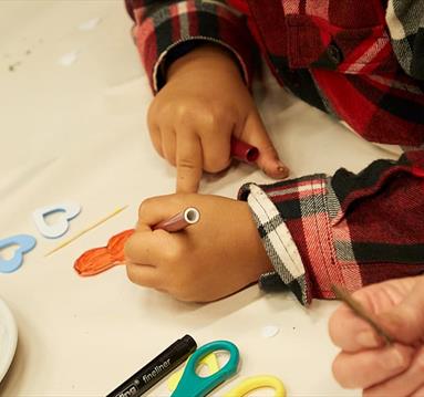 The image shows a child's hands, they are colouring a picture on a large piece of paper with an orange felt tip pen.