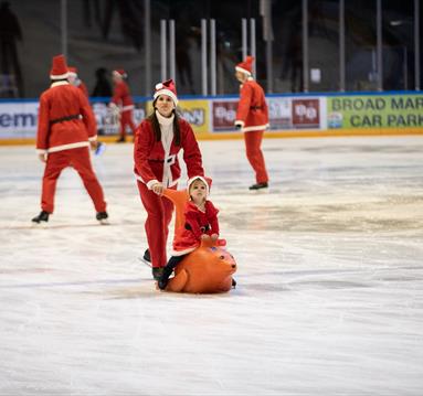 Photo of a woman and her child skating on the ice. They are both dressed as Santa