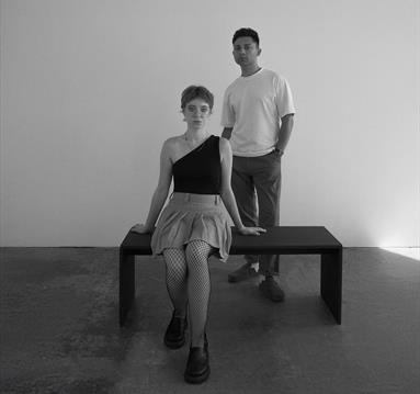 Image of a lady sat on a bench with a young man stood behind. 