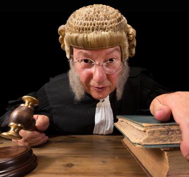 Photo of a person dressed as a judge leaning over their desk, gavel in hand, looking straight into the camera.