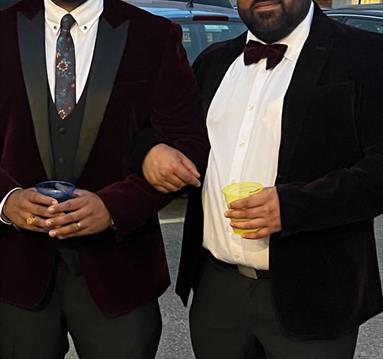 The image shows Tharun Chelley & Hitul Unadkhat stood in black tie suits holding a drink each and smiling into the camera.