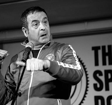 Image shows a black and white image of Mark Thomas on stage during his show - Gaffa Tapes.