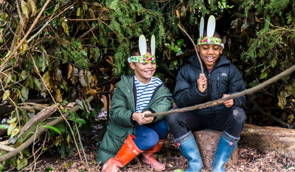 A photo of two young boys sitting on a log. They are smiling as they play with long twigs, and they are wearing rabbit ears attached to a paper crown.