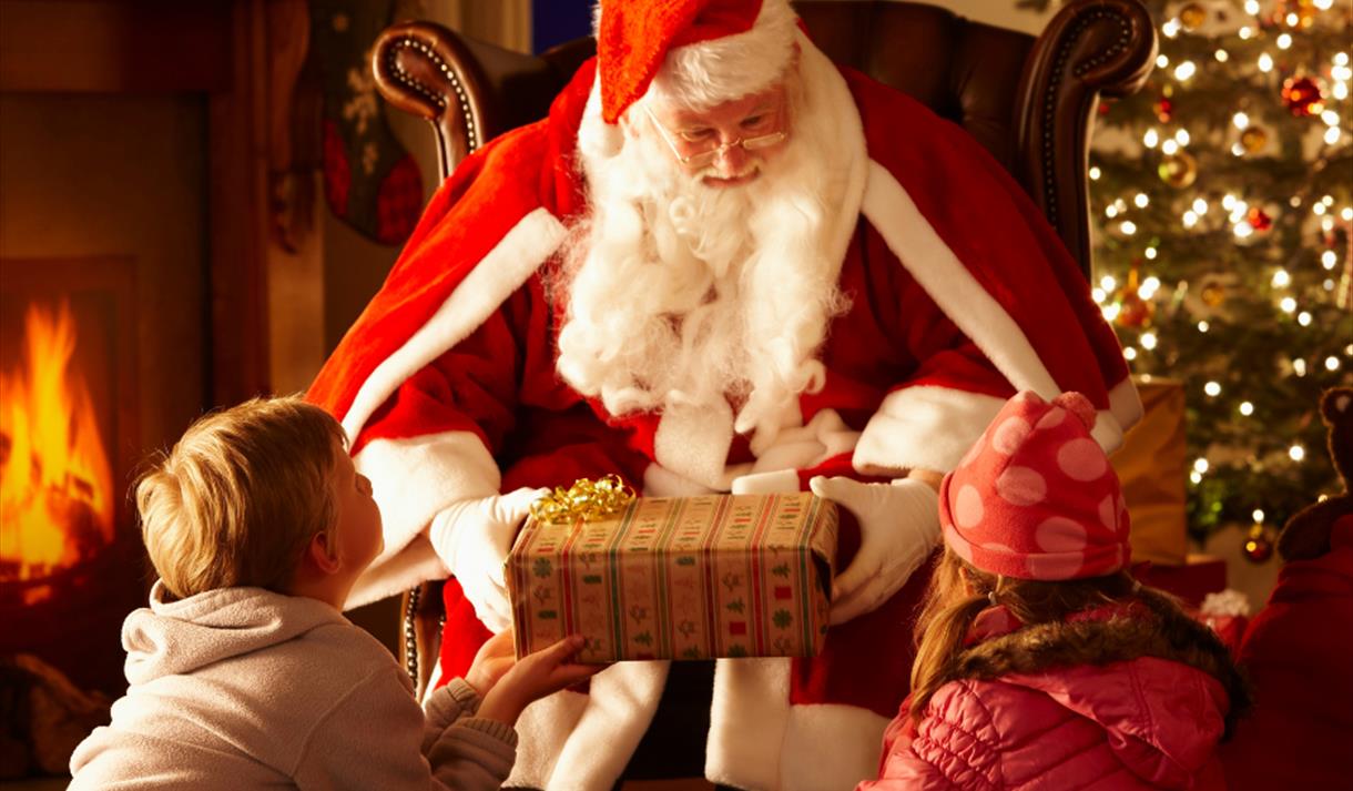 Two children being given a present by Santa in front of a Christmas tree and log fire