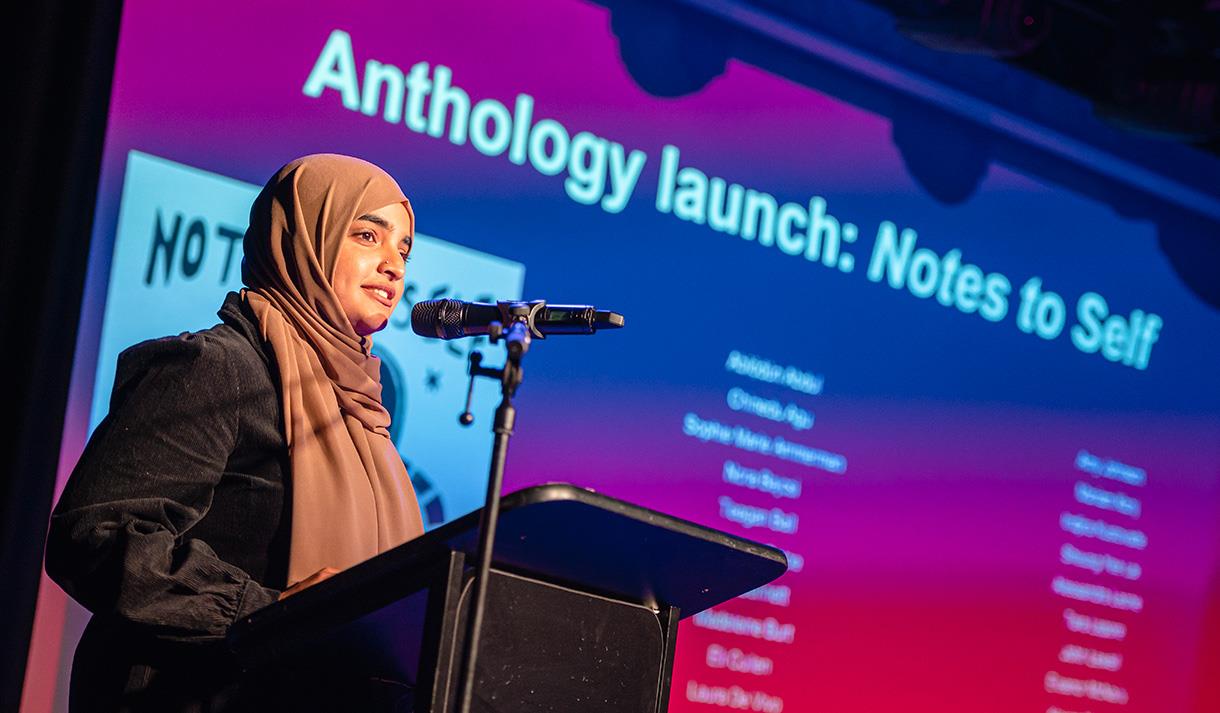 Image shows a girl on stage speaking into a microphone with a screen behind her reading - 'Anthology Launch: Notes to self.