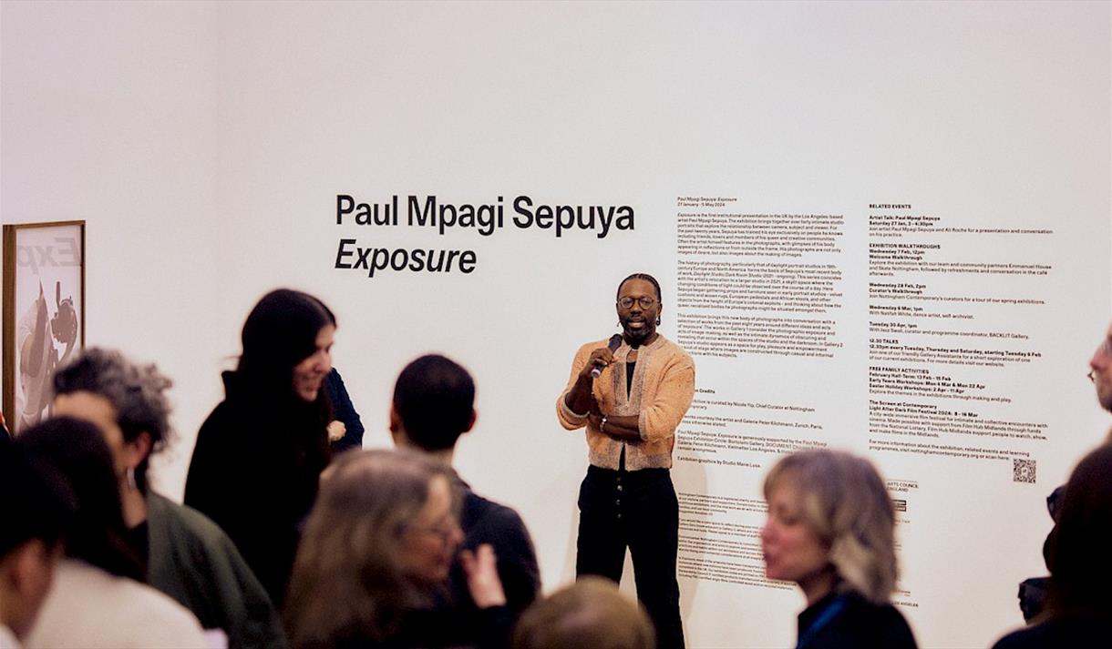 Photo of Paul Mpagi Sepuya on a walkthrough event. He is holding a microphone and you can see his gallery at Nottingham Contemporary in the background