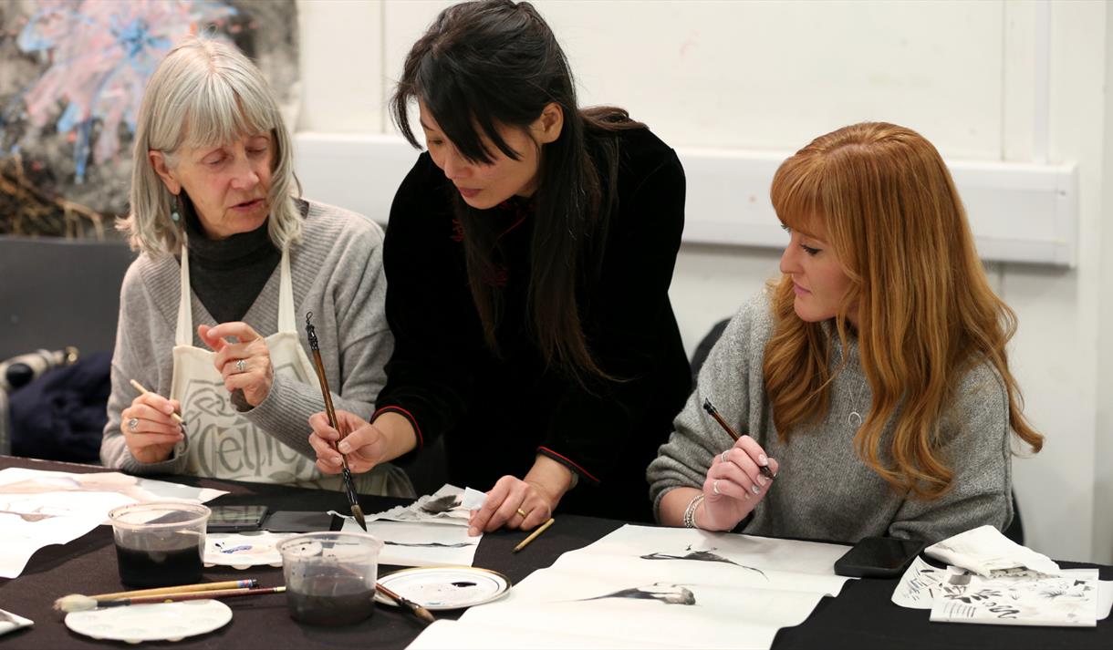 Photo of Feng-Ru Lee during a workshop. She is leaning over and assisting two women, who are holding paint brushes.