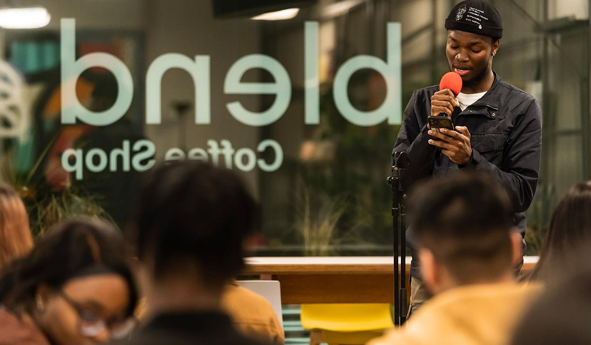 Image shows blend cafe sign in the background with a young boy holding an orange microphone wearing a black jacket, white top and black hat. There is