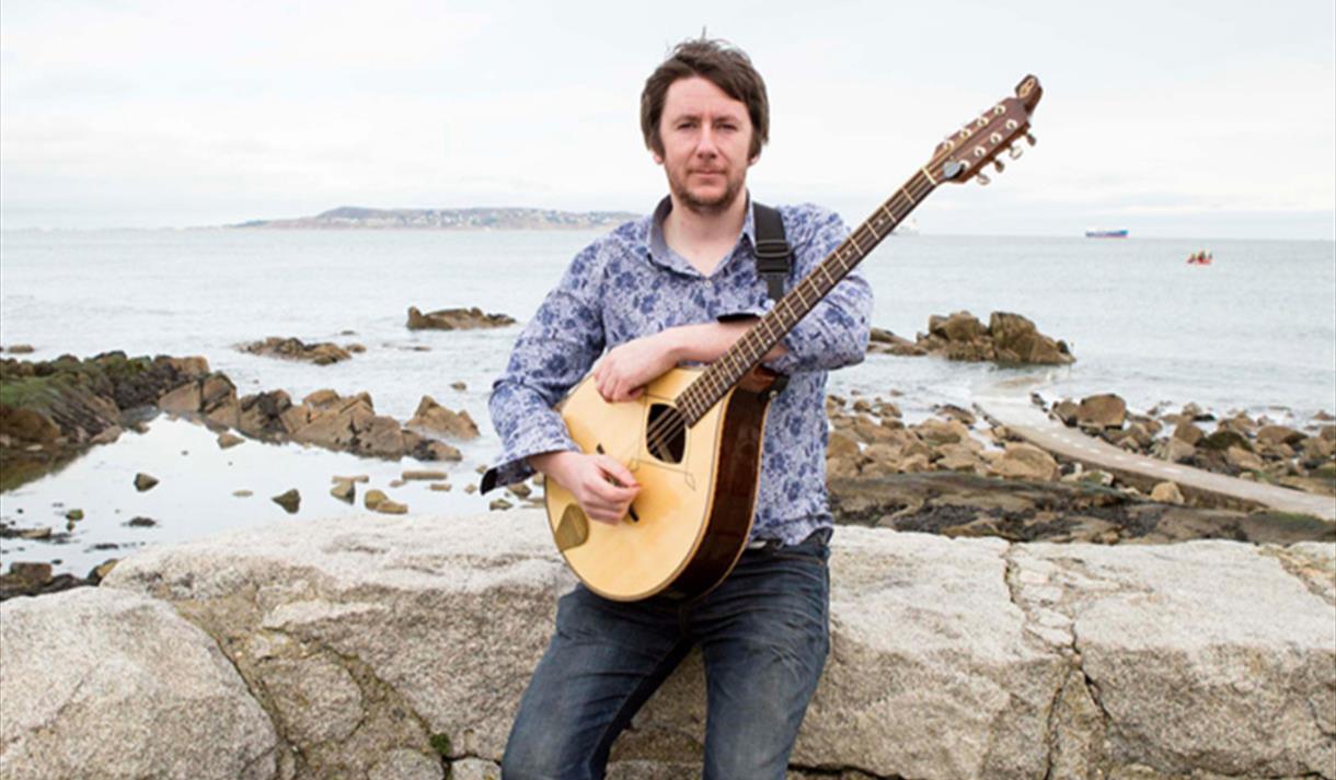 Irish singer and bouzouki player Daoirí Farrell sitting on a stone wall in front of a coastal scene