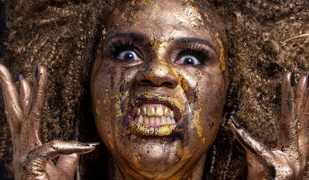 Desiree Burch covered in gold paint pulling a somewhat manic expression.