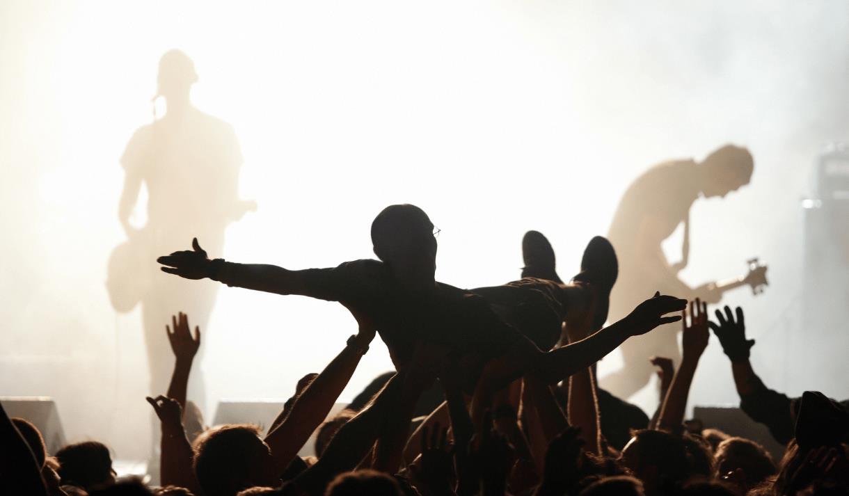 Photo of crowd surfer. They are in silhouette against bright white stage lights in the background. You can see some performers on stage.