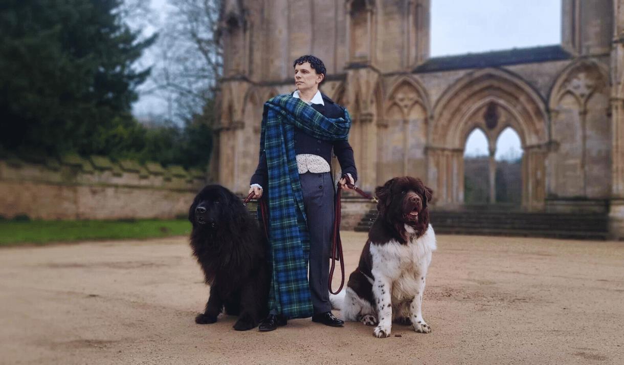 An actor dressed as Lord Byron poses in front of Newstead Abbey with two large Newfoundland dogs.