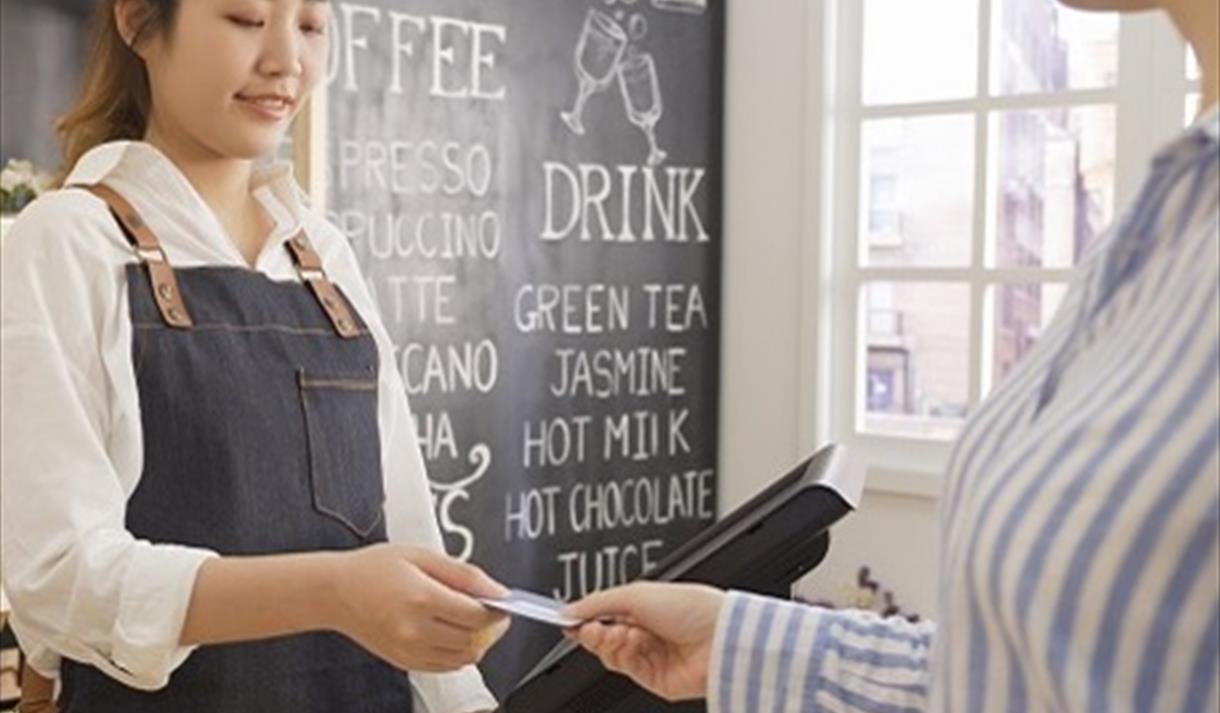 The image shows a lady behind a till taking something from someone's hand on the other side.