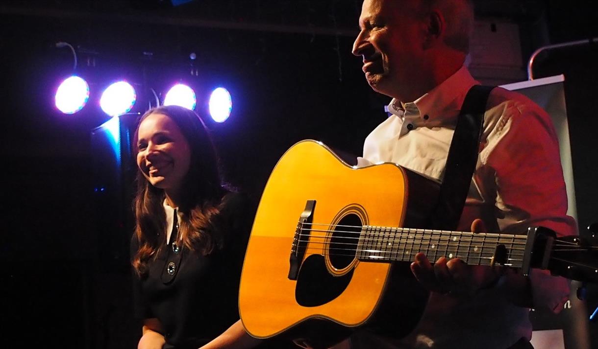 Image shows a man stood on stage playing a guitar and a woman stood next to him smiling.