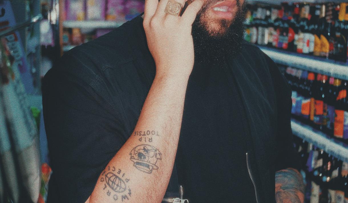 Jamali Maddix: Aston - Image shows a man on the phone stood in a supermarket wearing a black jumper and t shirt and holding two bottles of alcohol.