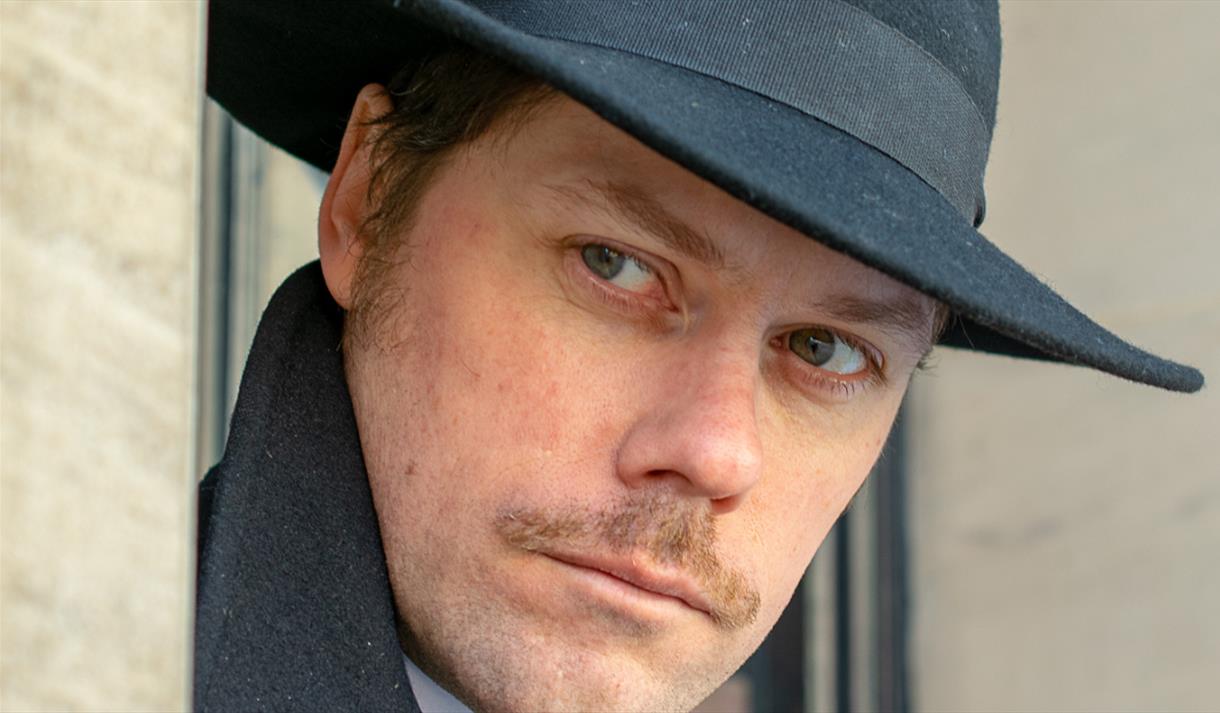 Man with fedora hat, pencil moustache and sinister appearance looks around a corner