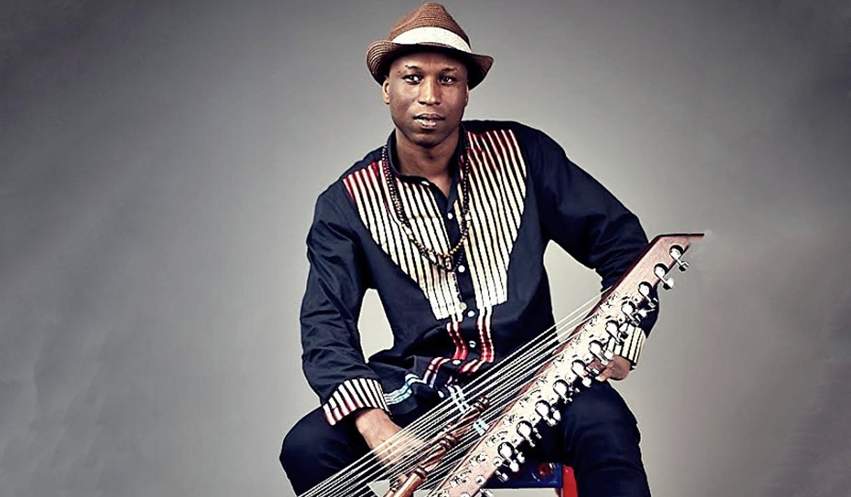 Image shows a man wearing a dark blue top with black and white stripes sat on a chair and holding an instrument.