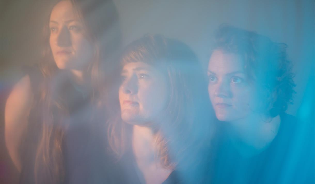 The image shows three women looking into the distance behind blue smoke.