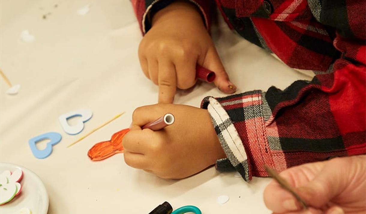 The image shows a child's hands, they are colouring a picture on a large piece of paper with an orange felt tip pen.