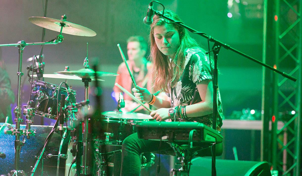 Image shows a girl on stage playing the drums with a microphone next to her.