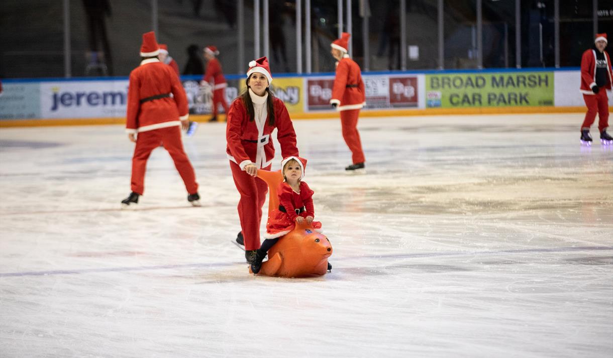 Photo of a woman and her child skating on the ice. They are both dressed as Santa