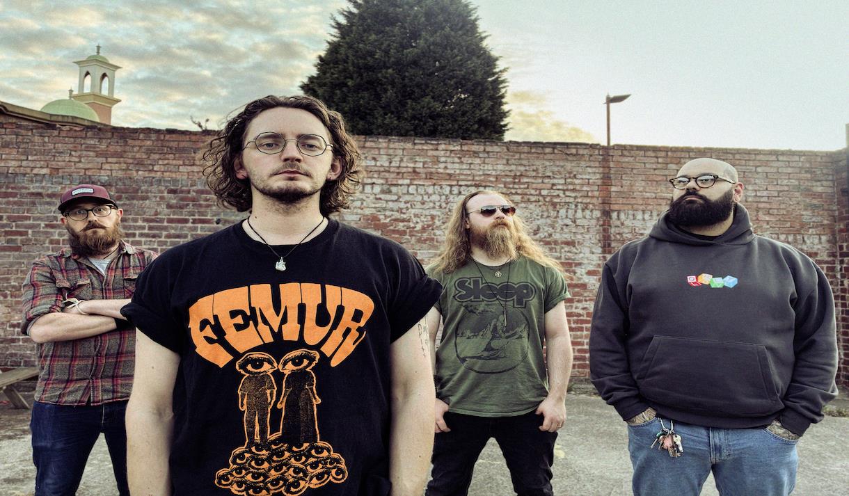 Four band members stand in front of a brick wall