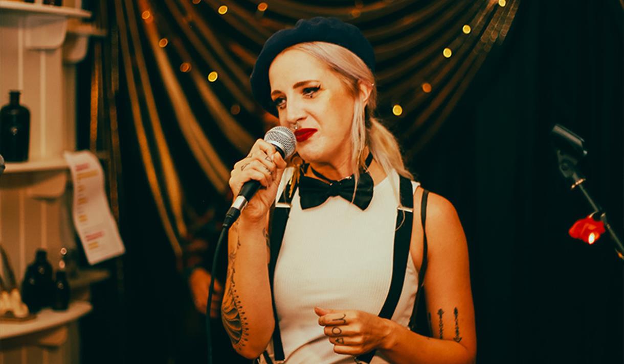 Image of young lady with blonde hair wearing a beret, bow tie and braces performing a spoken word piece on stage.