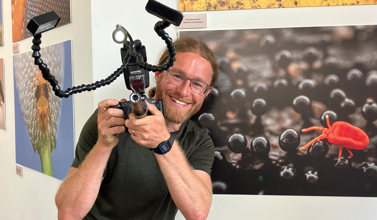 Alex Hyde holding a camera and smiling, with the sherwood forest up close exhibition wall behind