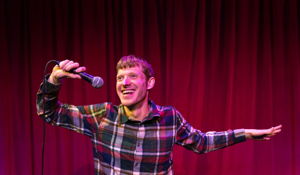 The image shows David Eagle stood on stage holding a microphone.