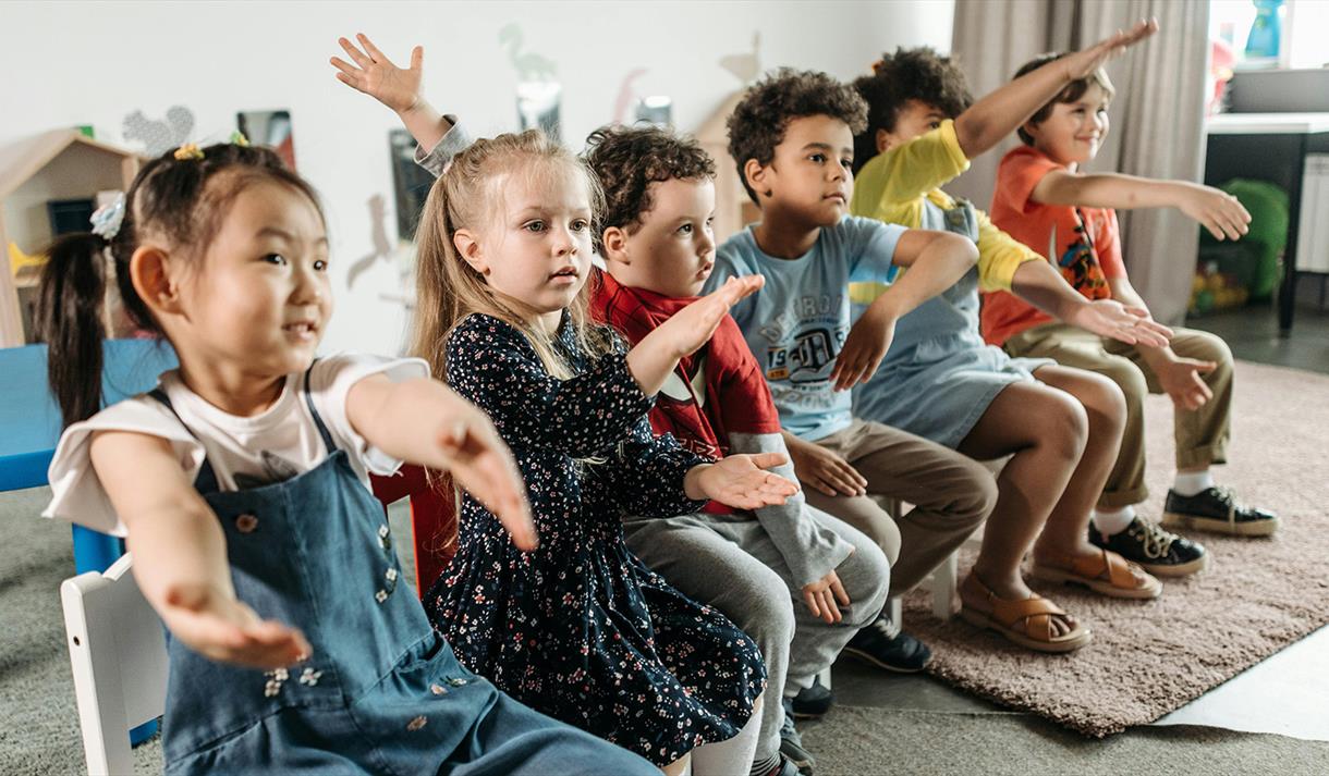 Photo of children sitting in a line of chairs.