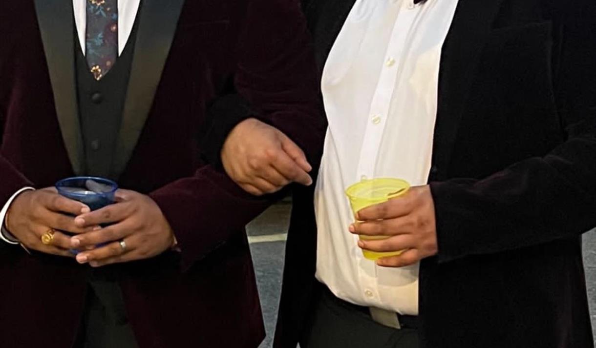 The image shows Tharun Chelley & Hitul Unadkhat stood in black tie suits holding a drink each and smiling into the camera.