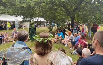 A crowd sits in a circle on a field for the Sherwood Festival. They sit engaged in a story by a cosumted figure.