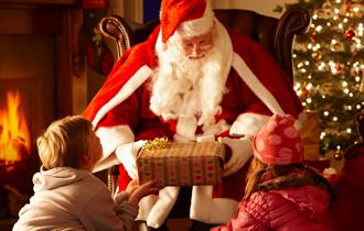 Two children being given a present by Santa in front of a Christmas tree and log fire