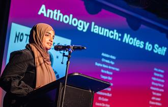 Image shows a girl on stage speaking into a microphone with a screen behind her reading - 'Anthology Launch: Notes to self.