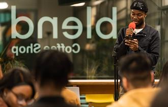 Image shows blend cafe sign in the background with a young boy holding an orange microphone wearing a black jacket, white top and black hat. There is
