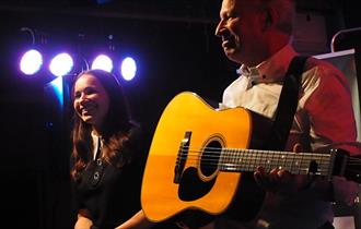 Image shows a man stood on stage playing a guitar and a woman stood next to him smiling.