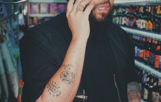 Jamali Maddix: Aston - Image shows a man on the phone stood in a supermarket wearing a black jumper and t shirt and holding two bottles of alcohol.